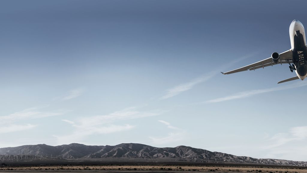 A Delta airplane airplane is flying upwards with its landing gear retracting, against a backdrop of rugged mountains.