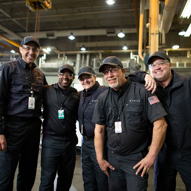 Four Delta mechanics in dark uniforms standing together in an industrial setting.
