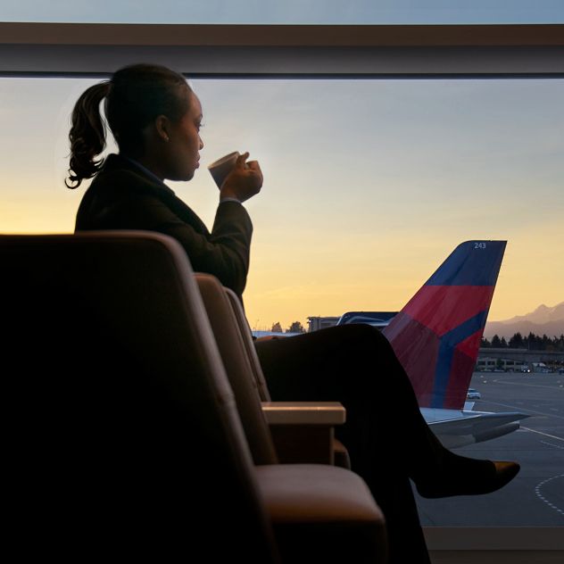 A person sitting in an airport lounge, looking out of a large window onto the tarmac. In the background, there is a mountain visible under the sunset sky.