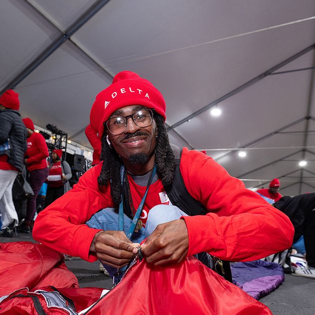 A person in a red shirt and a red DELTA hat sits and ties a knot on a red bag.