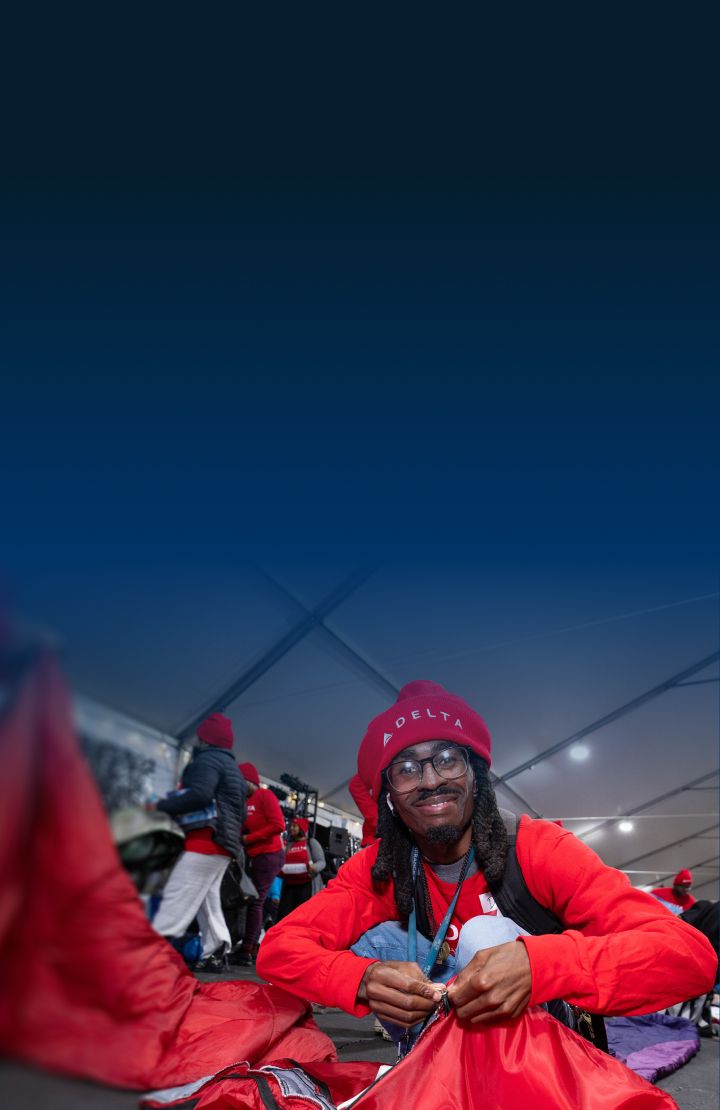 A person in a red shirt and a red DELTA hat sits and ties a knot on a red bag.