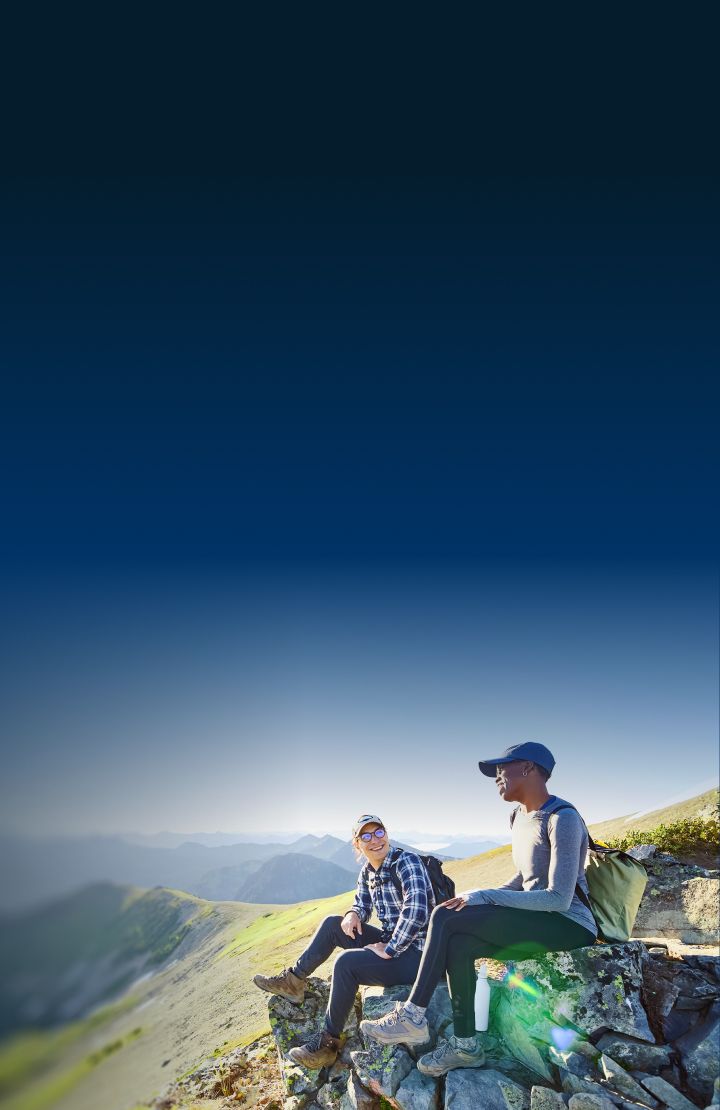 Two people sitting on a rocky ledge overlooking a mountainous landscape. The sky is clear and bright, and the terrain below includes green valleys and distant mountain ranges.