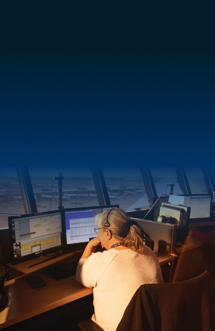 A woman sits at a desk with two computer monitors in an air traffic control tower.