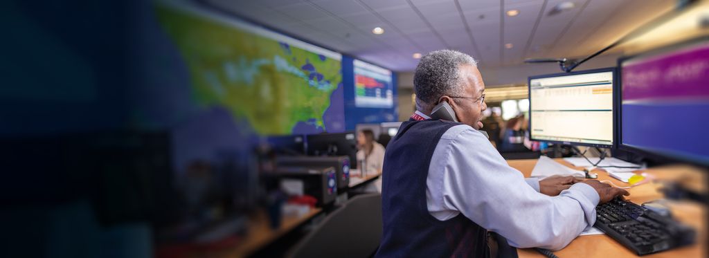 A Delta employee works at a desk with two computer monitors in a control room, wearing a white shirt and dark vest.