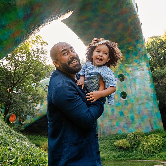 A man holding a child outdoors with greenery and a colorful structure in the background.