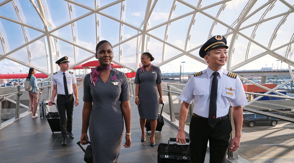 Delta pilots and flight attendants in uniform walk through an airport terminal with a modern glass ceiling, pulling rolling suitcases behind them.