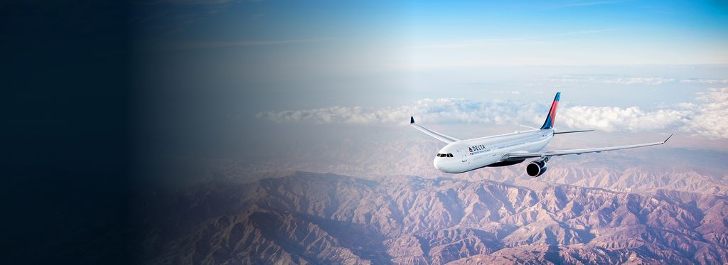 A Delta airplane flies over a vast, mountainous landscape with rugged terrain with clear skies and some distant clouds.