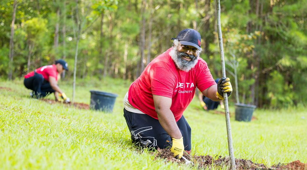 A Delta employee volunteer is planting a tree in a grassy area.