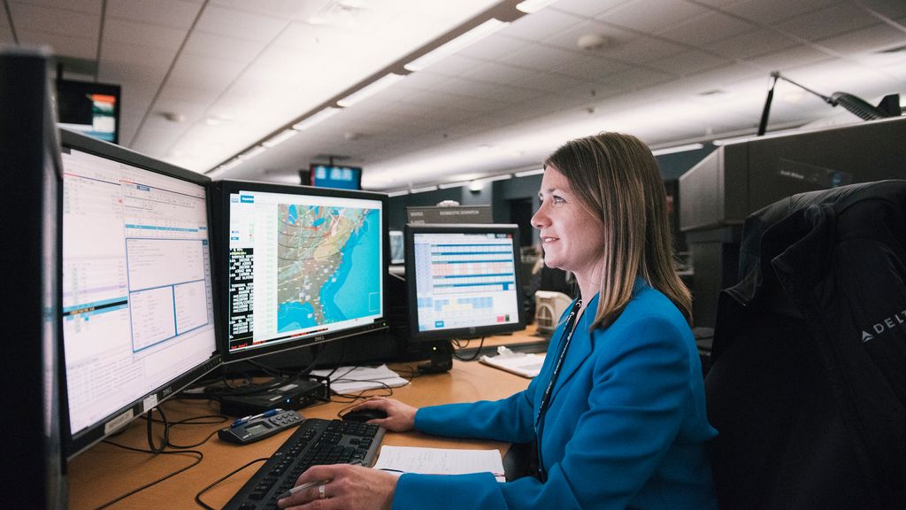 A person sitting at a desk in a control room with multiple computer monitors displaying various information, including maps and data.