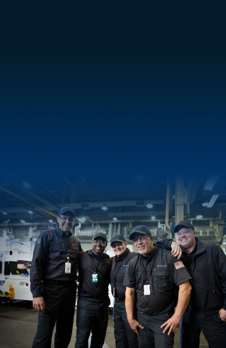 Four Delta mechanics in dark uniforms standing together in an industrial setting.