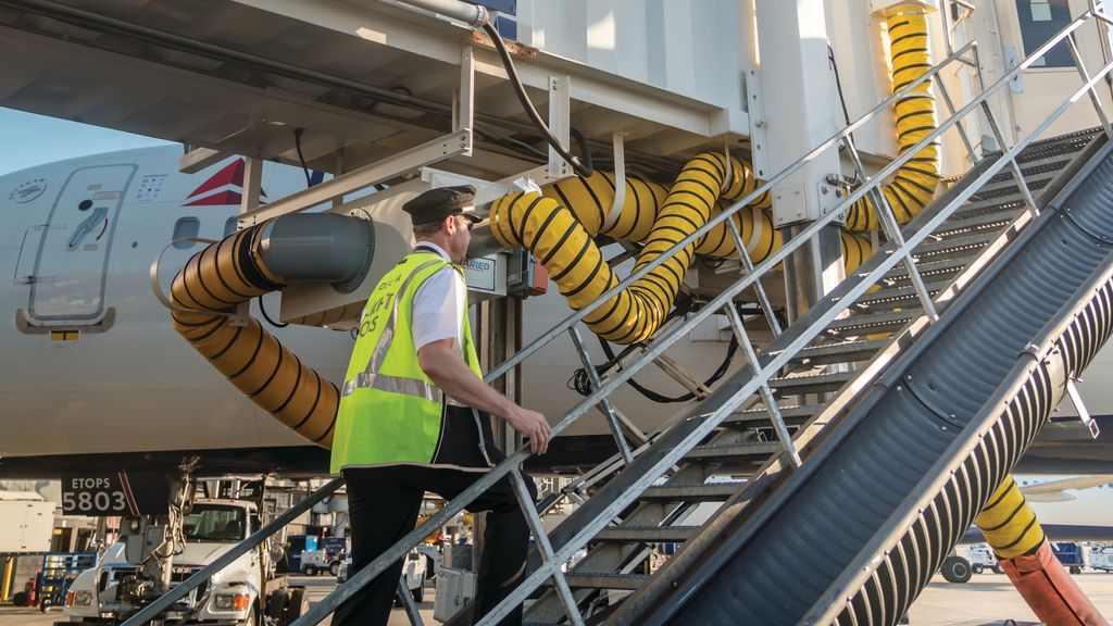 A pilot in a high-visibility vest walking up the jet bridge stairs towards a Delta airplane at an airport.