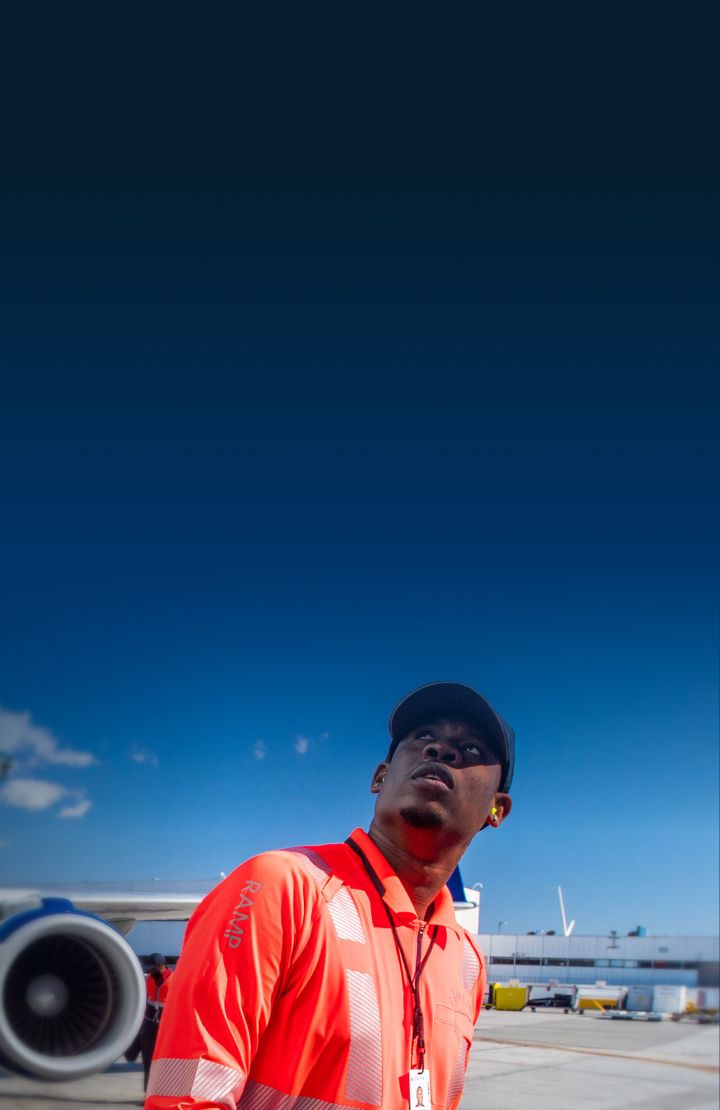 A Delta ground employee in an orange high-visibility vest holding two orange marshalling wands walks near a large airplane on an the tarmac.