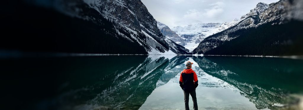 A person standing by a calm lake reflecting snow-capped mountains.