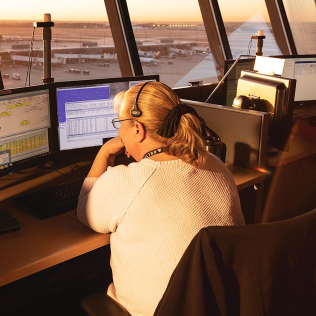 A woman sits at a desk with two computer monitors in an air traffic control tower.
