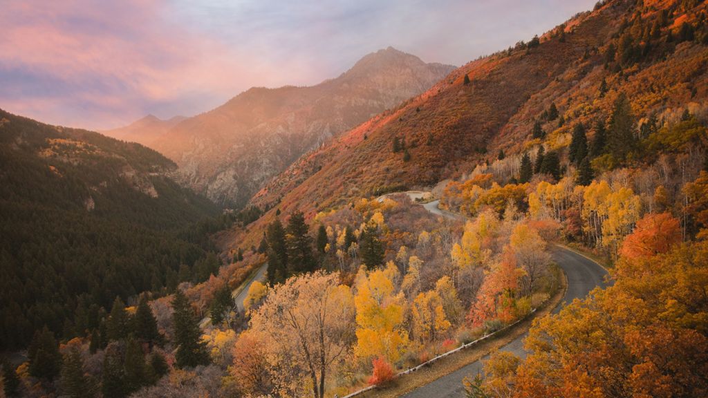 A scenic landscape with a woman running along a winding road surrounded by mountains covered with autumn foliage in vibrant shades of orange, yellow, and red.