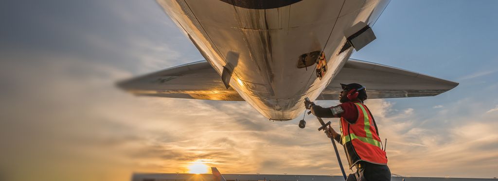 A Delta ground operations employee in an orange high-visibility vest holds a hose that connects to the underside of a Delta aircraft on the tarmac at sunset.