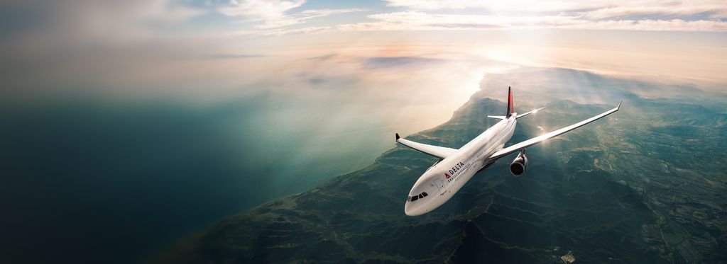 A Delta airplane viewed from above flies under a hazy sky above a rugged coastline and mountainous landscape. Sunlight reflects off the aircraft’s wings.