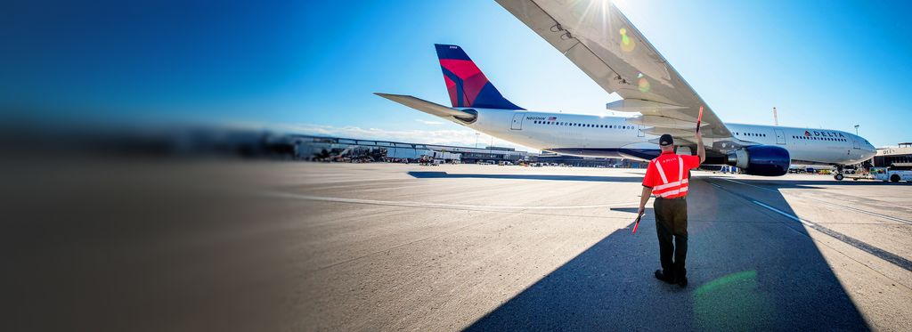 An airport ramp scene shows a ground crew member wearing an orange high‑visibility jacket standing on the tarmac and signaling with a handheld baton to a Delta airplane, assisting it as it parks at the gate under a clear blue sky.