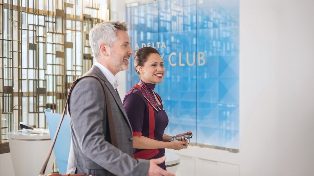 A Delta employee in uniform walks with a customer outside a Delta Sky Club lounge in an airport concourse.