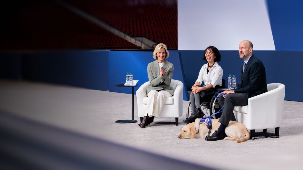 Three speakers sit on stage during a panel discussion. One person sits in a wheelchair at center, while two others sit in white chairs on either side. A small table with water bottles is positioned nearby against a blue and white backdrop.