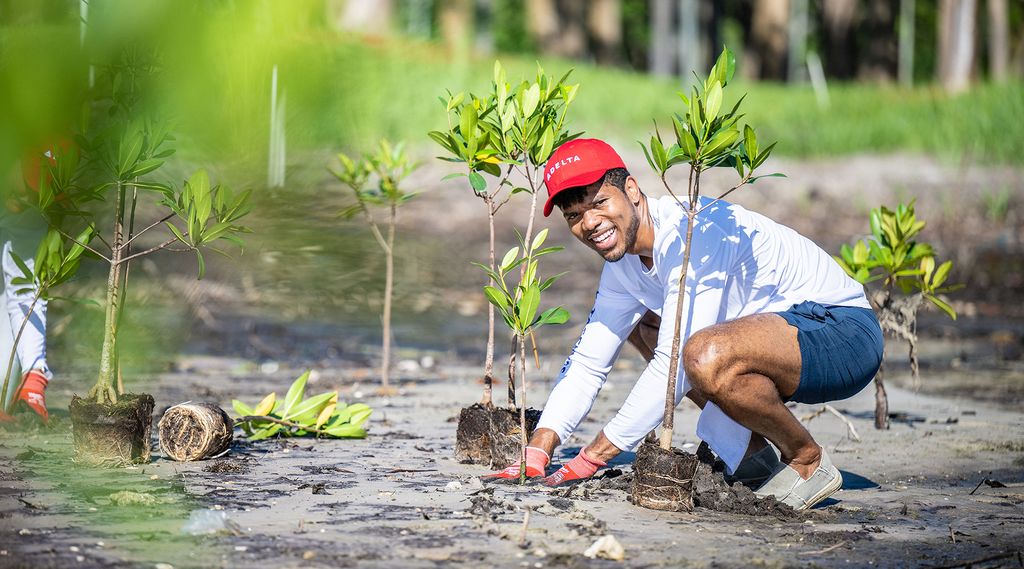 A person wearing a red cap, white long‑sleeve shirt, and blue shorts crouches outdoors to plant a small sapling in sandy soil. Several young plants are already placed nearby, with greenery and water visible in the background.