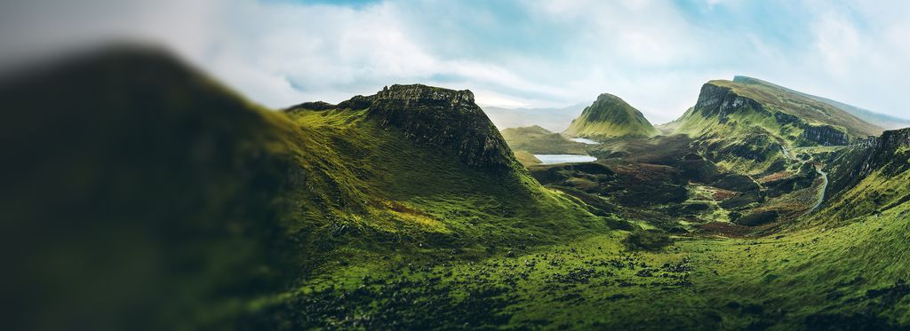 A wide view of a lush green valley with rolling hills, winding paths, and steep mountains rising in the background. Patches of sunlight highlight the landscape under a pale sky, creating a panoramic, natural scene.