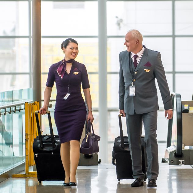 Two Delta employees in uniform each pulling a rolling suitcase walk side by side through a bright airport terminal, with large windows and an escalator visible in the background.