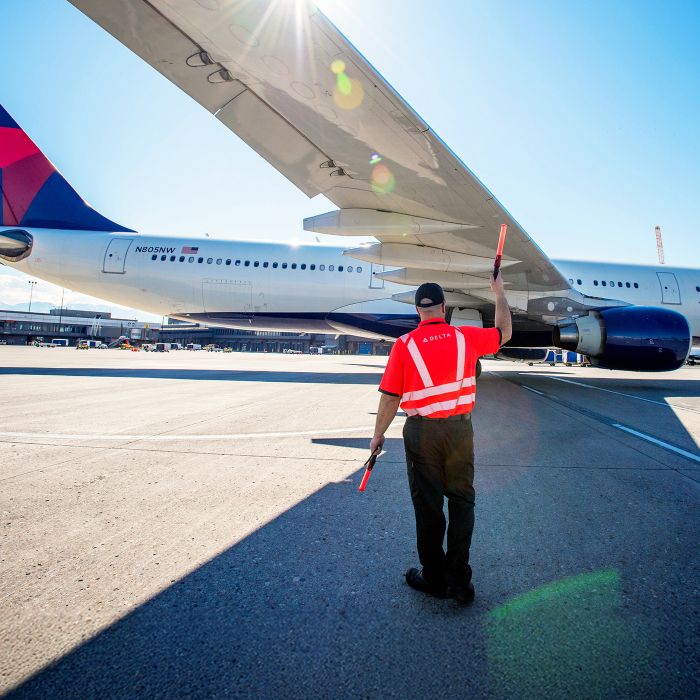 An airport ramp scene shows a ground crew member wearing an orange high‑visibility jacket standing on the tarmac and signaling with a handheld baton to a Delta airplane, assisting it as it parks at the gate under a clear blue sky.