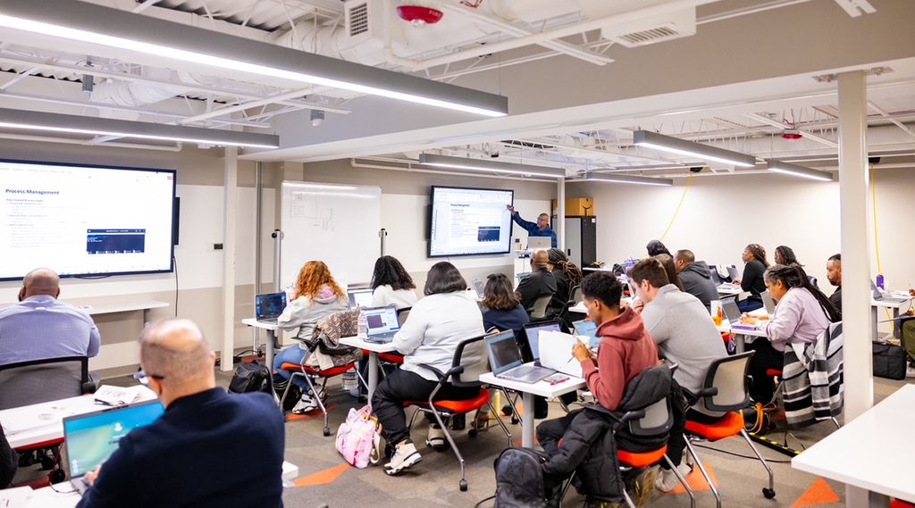 A classroom setting shows a group of adults seated at tables, working on laptops and facing presentation screens at the front of the room.