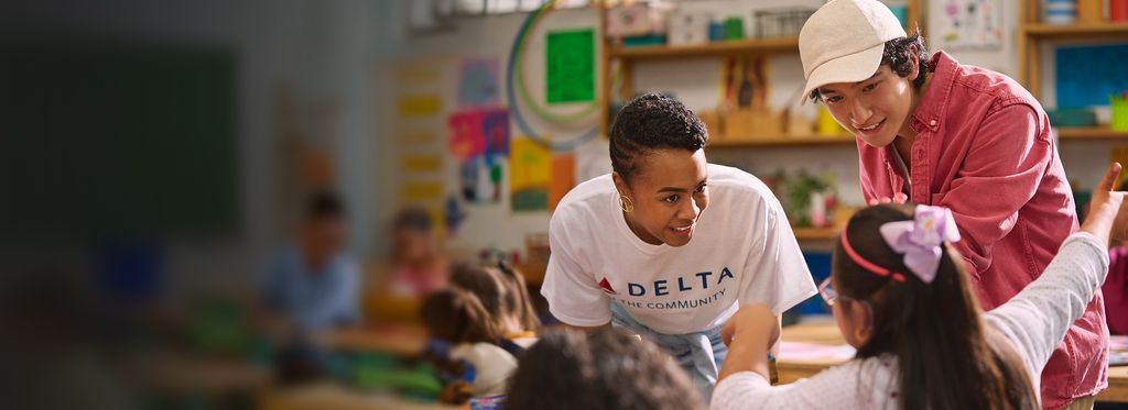 Inside a classroom, a Delta employee wearing a white “DELTA” T‑shirt leans forward to assist a child seated at a table, while another adult in a pink jacket and white hat stands nearby. Children’s artwork and classroom materials fill the background.