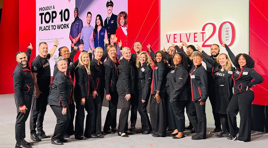 A group of smiling Delta employees dressed in coordinated black attire with red accents poses together on a stage at an event, in front of a red backdrop that reads “Top 10 Places to Work” and “Velvet 20 years.” 