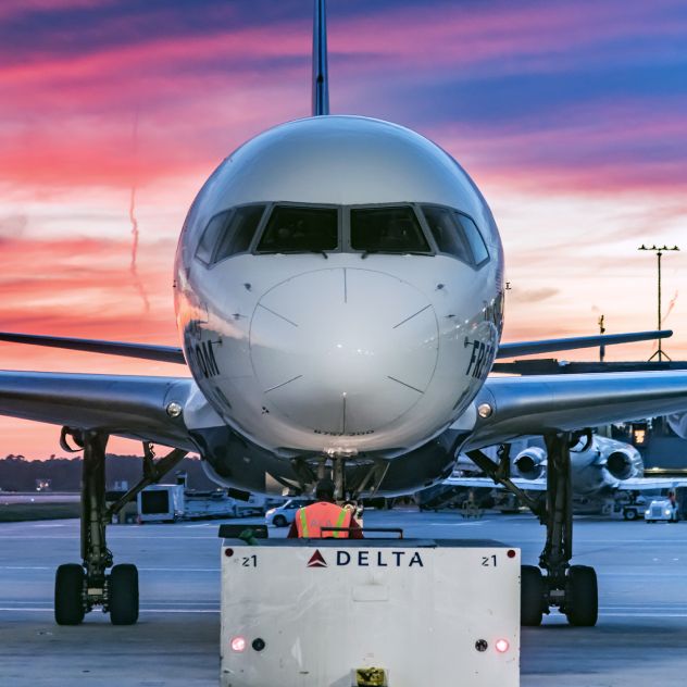 A close up, front-facing  view of a Delta airplane on the tarmac at sunset, with an employee in an orange high‑visibility vest standing directly in front of the nose and facing the aircraft. Pink and orange clouds fill the sky, with airport equipment visible beneath the wings.
