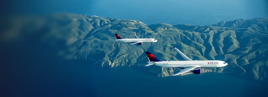 Two Delta airplanes fly side by side above open water, with a lush mountain range visible in the distance under a clear sky.