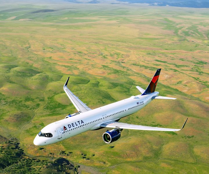 A Delta airplane flies above a green landscape of fields and rolling terrain, viewed from an elevated angle, with the aircraft’s white body and red‑and‑blue tail clearly visible
