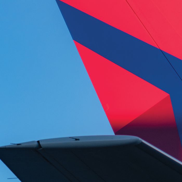 The tail section of a Delta airplane, featuring the red and blue Delta widget logo, extends over a tarmac, with a ground crew member standing below against a clear blue sky.
