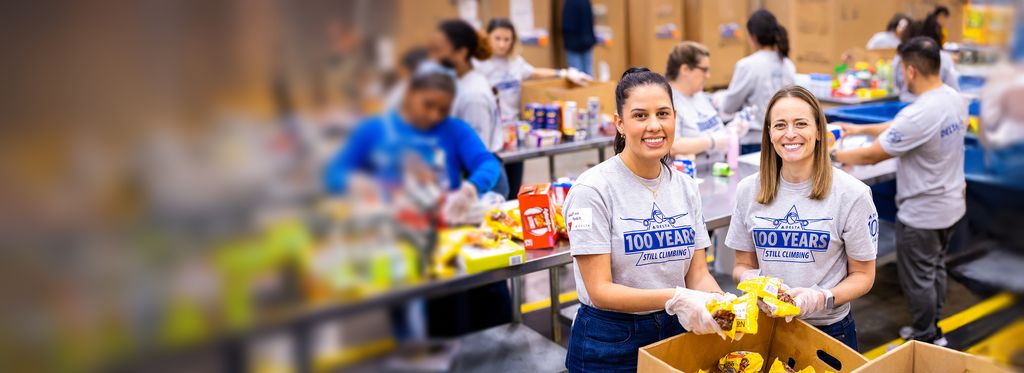 Two Delta employees stand side by side wearing matching gray T‑shirts that read “100 YEARS – STILL CLIMBING.” They hold packaged food items, with tables, boxes, and other volunteers working in the background