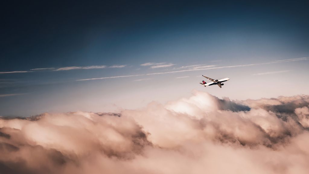 A Delta airplane climbs through the sky above soft clouds, viewed from below, with warm light illuminating the aircraft against a pale blue and pink horizon.