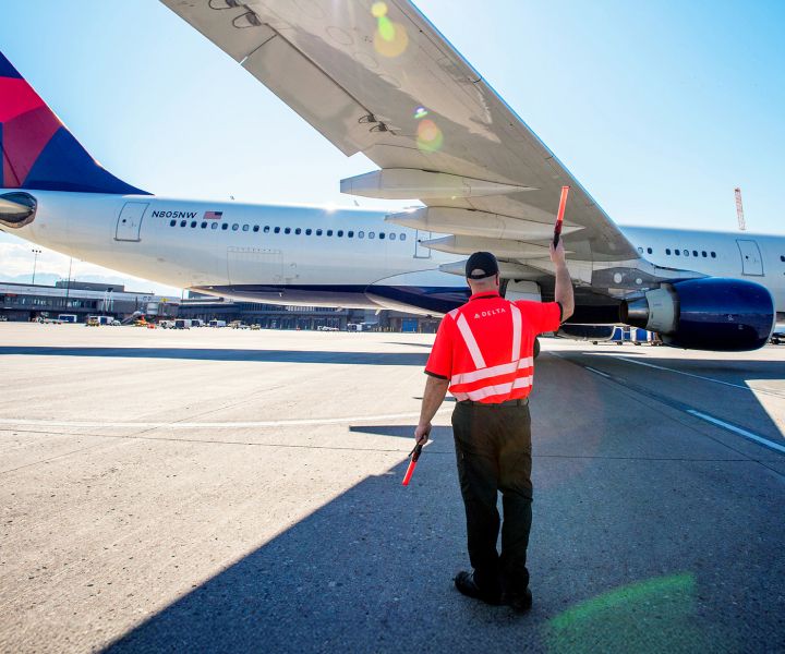 An airport ramp scene shows a ground crew member wearing an orange high‑visibility jacket standing on the tarmac and signaling with a handheld baton to a Delta airplane, assisting it as it parks at the gate under a clear blue sky.