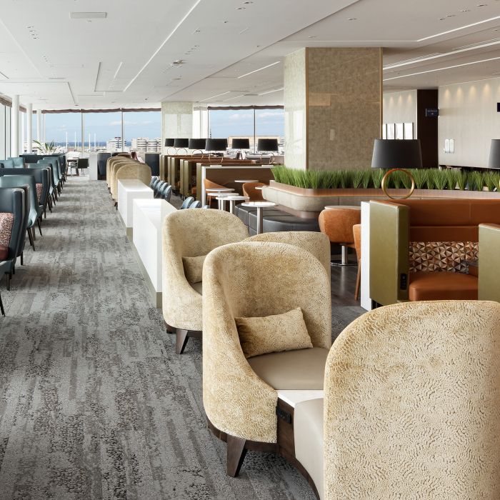 Rows of upholstered lounge chairs and sofas arranged along a carpeted Delta Sky Club lounge, with low tables and planters separating seating areas.