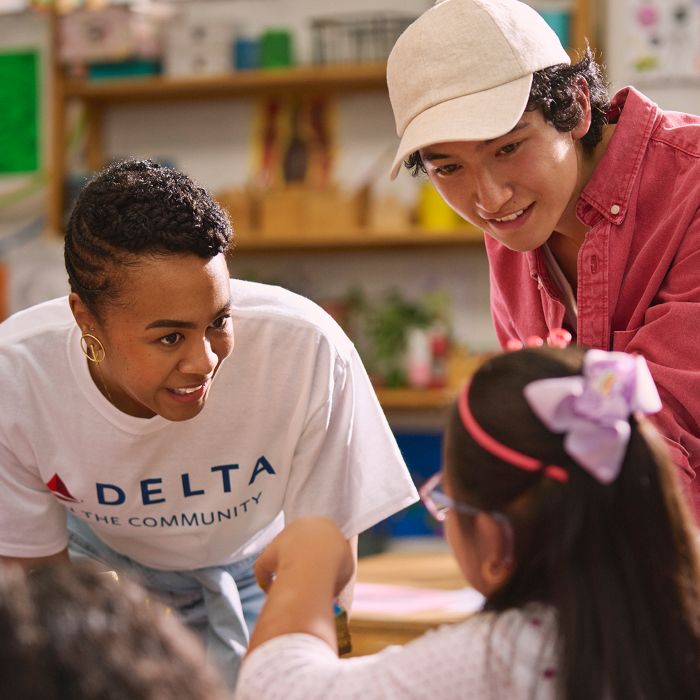 Inside a classroom, a Delta employee wearing a white “DELTA” T‑shirt leans forward to assist a child seated at a table, while another adult in a pink jacket and white hat stands nearby. Children’s artwork and classroom materials fill the background.