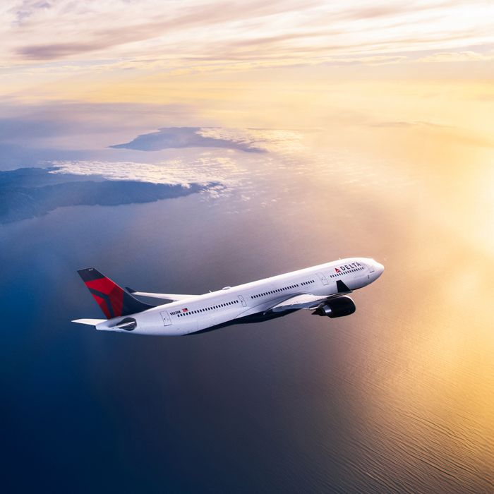 A Delta airplane flies above a sunlit ocean at golden hour, viewed from the side with warm light reflecting off the fuselage and wings against a glowing sky.