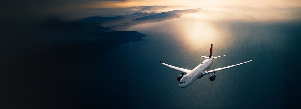 A Delta airplane flies above open water, viewed from above, with sunlight reflecting off the wings and the ocean below glowing in warm light.