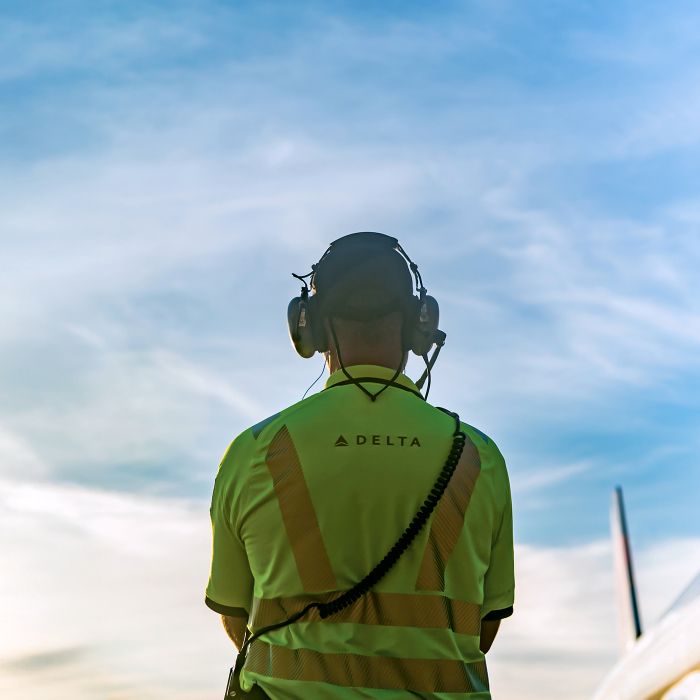 A ground operations employee seen from behind wearing a high‑visibility yellow Delta shirt and a headset, standing outdoors and facing the airfield under a blue sky.