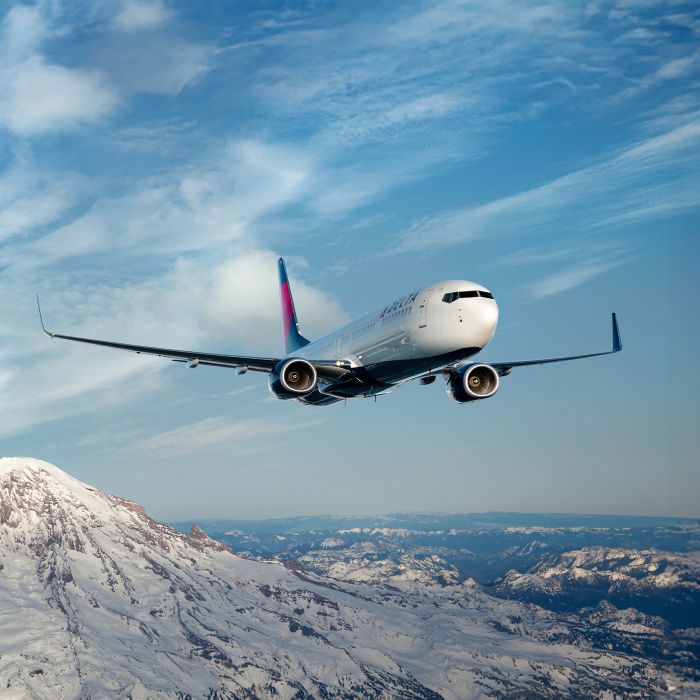 A Delta airplane flies above snow‑covered mountains under a clear blue sky.