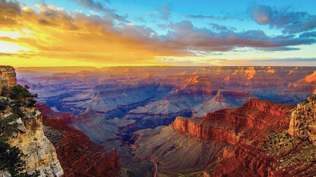 A wide canyon at sunset, with layered red rock formations glowing in golden light beneath a blue sky with scattered clouds.