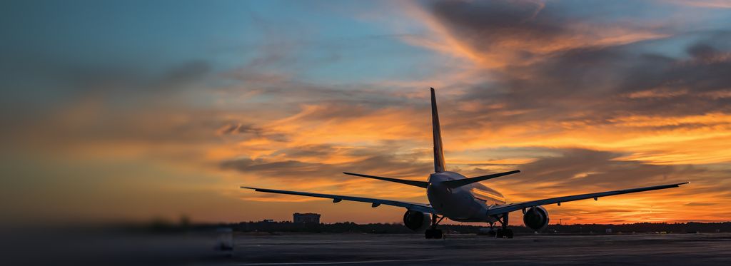 A Delta airplane is seen from behind on a tarmac, silhouetted against a dramatic orange and gold sunset sky.