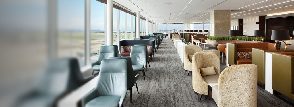 Rows of upholstered lounge chairs and sofas arranged along a carpeted Delta Sky Club lounge, with low tables and planters separating seating areas.
