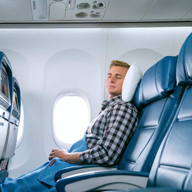 Inside an airplane cabin, a passenger wearing a black-and-white checkered shirt rests in a blue leather window seat with eyes closed, head on a white pillow and legs covered with a blue blanket, as daylight comes through the window