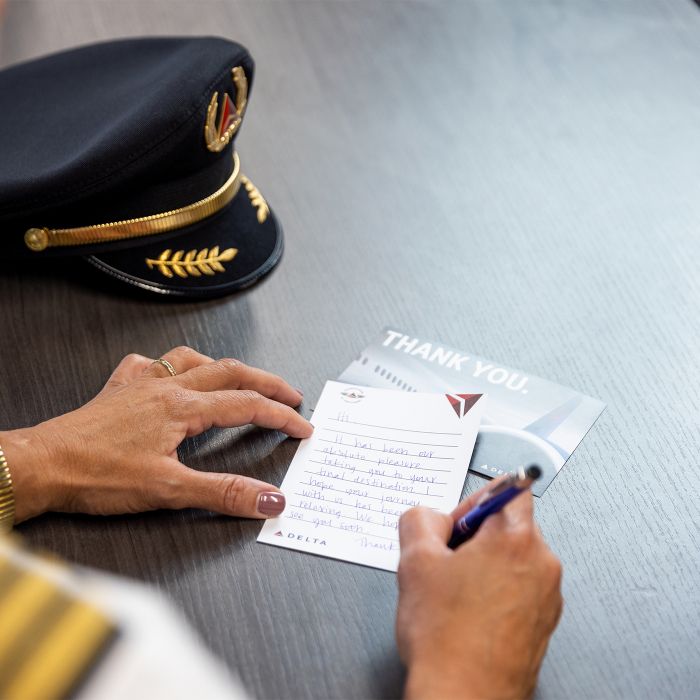 Close-up view of a uniformed Delta pilot seated at a desk, writing a handwritten note on a Delta-branded “Thank You” card. A pilot’s hat rests on the desk beside the card. The pilot’s sleeve with striped insignia is visible, emphasizing a professional aviation setting and a gesture of appreciation.