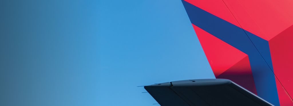 The tail section of a Delta airplane, featuring the red and blue Delta widget logo, extends over a tarmac, with a ground crew member standing below against a clear blue sky.
